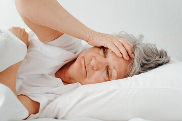 Fototapeta na wymiar Headache, insomnia, depression, women's health concept. Side view of lonely upset senior woman holding her head while lying in bed in bedroom