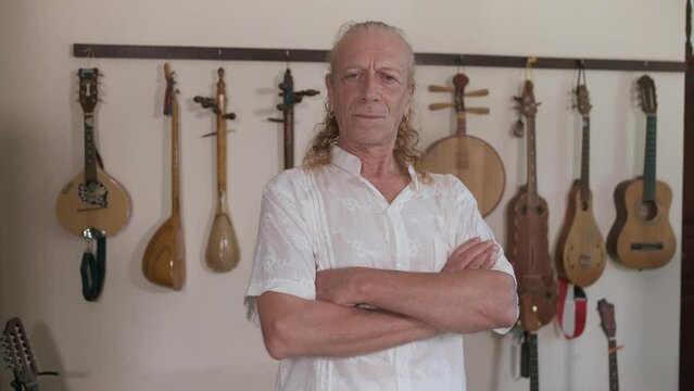 Elderly caucasian musician in white shirt stands in his home studio. Behind him on the wall is a collection of oriental stringed musical instruments.