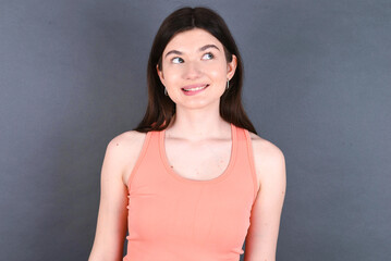 Amazed Caucasian woman wearing orange T-shirt over grey wall bitting lip and looking tricky to empty space.