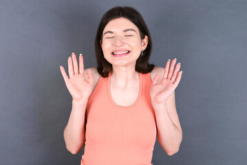 Emotive Caucasian woman wearing orange T-shirt over grey wall laughs loudly, hears funny joke or story, raises palms with satisfaction, being overjoyed amused by friend