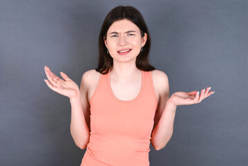 Clueless Caucasian woman wearing orange T-shirt over grey wall shrugs shoulders with hesitation, faces doubtful situation, spreads palms, Hard decision
