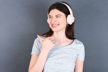  young beautiful Caucasian woman wearing white T-shirt over grey wall listens audio track via wireless headphones points thumb away advertises copy space