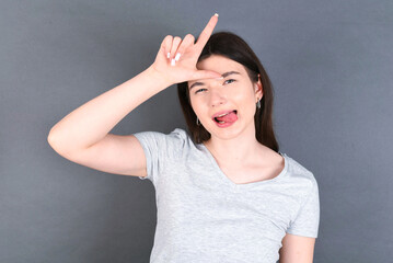 Funny young beautiful Caucasian woman wearing grey T-shirt over grey wall makes loser gesture...