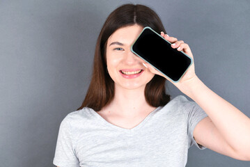 young beautiful Caucasian woman wearing white T-shirt over grey wall holding modern smartphone covering one eye while smiling