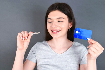Young beautiful caucasian girl standing over grey background holding a bank credit card and an invisible aligner braces and smiling. Dental healthcare concept. 