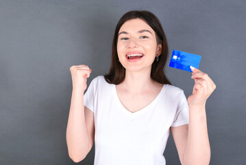 Young beautiful caucasian woman wearing white T-shirt over gray background holding a credit card, screaming and making winner gesture clenching and rising fist up.