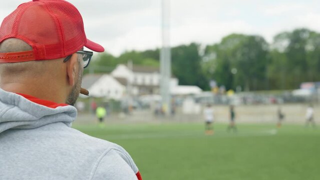 Football coach with red cap and glasses smoking cigar while watching his team during soccer match