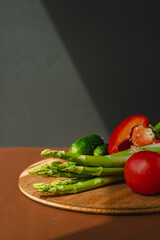 Vegetables lie on a wooden board: tomatoes, asparagus, cucumbers, red bell peppers. brown, dark gray background. place for text.