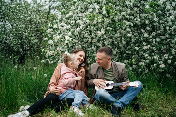 family dad mom baby daughter in the garden blooming apple trees, father playing the ukulele, scent of flowers outdoor nature