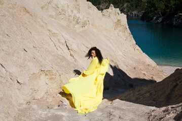 beautiful brunette woman with curly hair dancing in long yellow dress on the beach in mountains