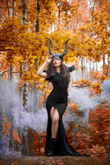 Naklejka premium Concept of Halloween and fantasy horror. Cosplay on Maleficent demonic - starring. Face of beautiful woman from a fairytale in autumn forest in the smoke. Beautiful girl dressed up as devil