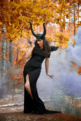 Naklejka premium Concept of Halloween and fantasy horror. Cosplay on Maleficent demonic - starring. Face of beautiful woman from a fairytale in autumn forest in the smoke. Beautiful girl dressed up as devil