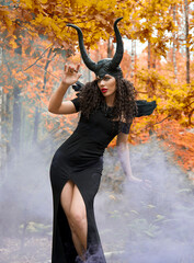 Naklejka premium Concept of Halloween and fantasy horror. Cosplay on Maleficent demonic - starring. Face of beautiful woman from a fairytale in autumn forest in the smoke. Beautiful girl dressed up as devil