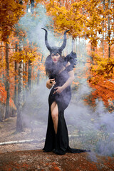 Concept of Halloween and fantasy horror. Cosplay on Maleficent demonic - starring. Face of beautiful woman from a fairytale in autumn forest in the smoke. Beautiful girl dressed up as devil