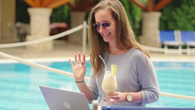 Woman on video chat from vacation. Using laptop computer and drinking cocktail on poolside, outdoor.