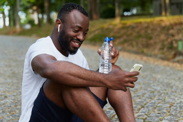 Young adult man resting on a park after a run with a bottle of water and smartphone. Outdoor workout