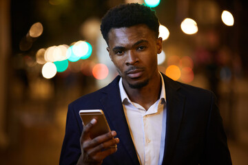 Portrait of serious handsome young black businessman with beard wearing suit and white shirt checking phone while walking over night city