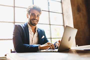 Happy young businessman typing on a laptop computer against a window - portrait of an entrepreneur...