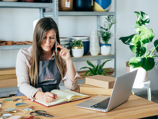 Smiling woman artisan speaking on her phone with client and writing his order in notebook while sitting at workshop desk, using laptop computer and packing merchandise for delivery in cardboard boxes