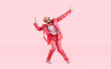 Fototapeta na wymiar Happy young dancer guy in pink suit dancing in the studio. Funny cheerful bald bearded man wearing a suit, bowtie, trainers and eyeglasses dancing and having fun isolated on pink color background