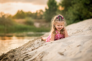 little girl is lying on the sand on the beach at the sunset and laughing 
