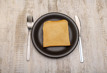 A single slice of toast on a plain black plate and cutlery with signs of use on a rustic wooden...