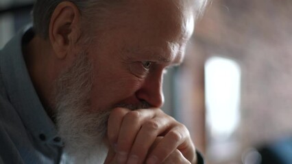 Close-up head shot of bearded gray-haired frustrated thoughtful senior adult man sitting alone at home thinking of lonely life. Sad depressed elderly male suffering from loneliness at nursing home. - Powered by Adobe