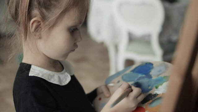 Close-up of cute little girl wearing black dress painting on a canvas with paintbrush and colourful acrylic paints, holding palette, children artist concept