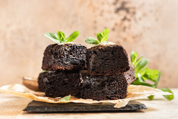 Stack of brownies made of dark chocolate with cocoa powder and mint on baking paper, brown background. Tasty fudge and chewy dessert with melting pieces of chocolate.