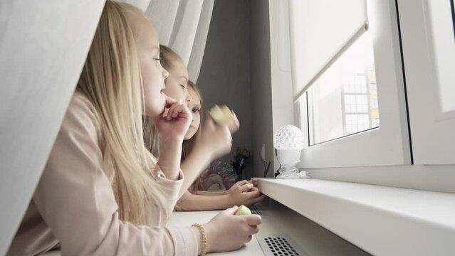 Close-up Of Three Little Girls Watching Out Of Window, Looking At The Street, Eating Apples And Laughing. Sisters Spending Time Together At Home. Family Togetherness Concept.