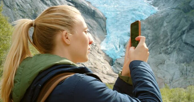 4k video footage of a young woman standing and using her cellphone to photography Briksdal Glacier