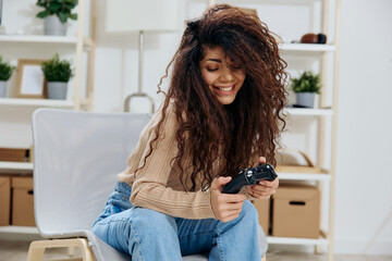 Laughing pretty enjoyed tanned curly Latin winner lady hold with joystick gamepad in both hands sit on chair in home modern interior look down. Copy space Mockup Banner. Concept game platform