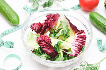 Lettuce leaves in a glass bowl with cucumbers, tomato, fresh dill and a measuring tape on a cement .