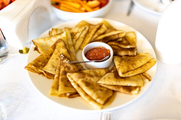 Pancakes folded in a triangle are on a plate around a bowl with red caviar and a spoon on a table in a restaurant on Shrove Tuesday