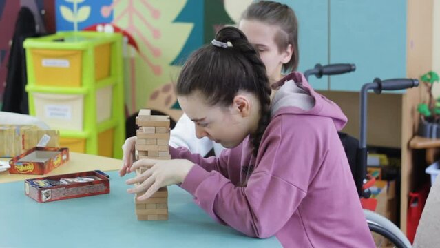 two girls with disabilities are playing a board game sitting at a large table. Rehabilitation of people with disabilities.