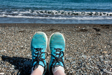 Fototapeta premium Women's legs in blue sneakers on a pebble beach against the backdrop of the sea. Sports and relaxation on the sea coast. Mediterranean Sea, Turkey, Antalya, Kemer.