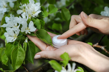 The moisturizing skincare cream in an open glass jar on woman's palm among the white flowers....