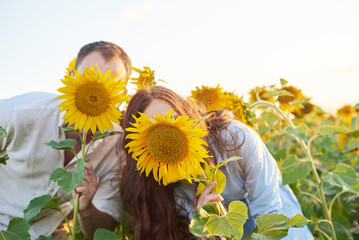 Funny loving couple of young women and men cover their faces with sunflowers in the field. Love in...