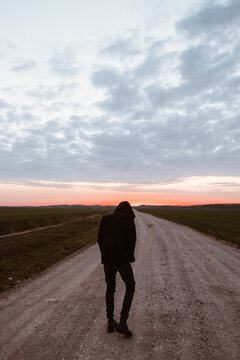 the guy in the hood with his back to the camera is walking along the road against the sunset in an open field