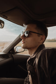 portrait of a guy in a shirt and sunglasses sitting in a car in the rays of sunlight in the field