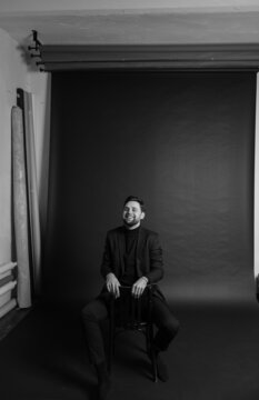 black and white photo of a guy in a stylish suit sitting on a chair and laughing looking at the camera in the studio