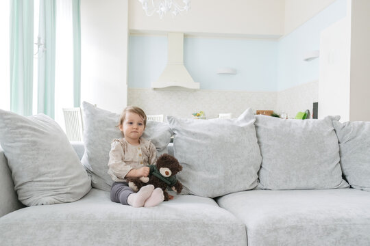 little girl in a bright apartment carefully looks forward with a bear in her arms