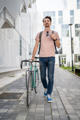 Fototapeta na wymiar One man young adult male with brown hair and mustaches walking by the building with rucksack on his back and bicycle happy smile joyful real people copy space full length front view