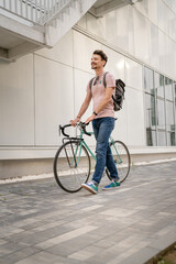 One man young adult male with brown hair and mustaches walking by the building with rucksack on his back and bicycle happy smile joyful real people copy space side view full length © Miljan Živković