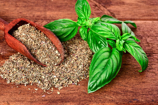 Crushed dried basil with wooden scoop and green leaves on brown wooden board. Selective focus