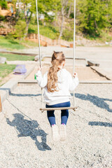 A little girl swings on a wooden swing in the park on the playground. Back view. Outdoor activities