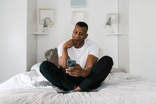 Young black guy biting apple and using smartphone