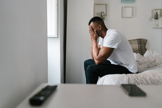 Tired black man covering face and sitting on bed