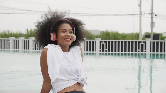 Young African American woman wearing headphones listening music at swimming pool, female wearing earphones while enjoy with happiness at poolside, emotion and feeling, lifestyles concept.