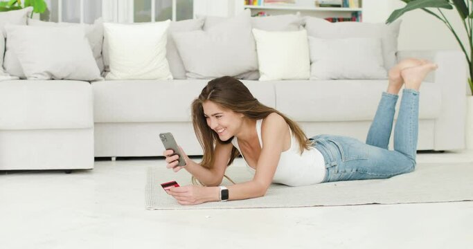 Young beautiful woman is lying on the floor in the living room making purchases via the Internet using a smartphone and a bank card. In the background is a white sofa. Modern technology concept.